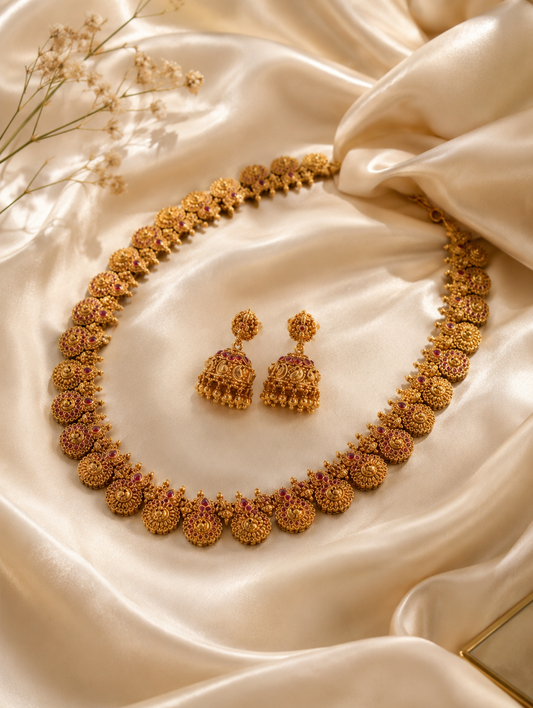 An antique gold-toned temple jewelry set featuring a long haram necklace made of continuous circular floral motifs accented with rubies, paired with matching hanging jhumkas on a black display bust.