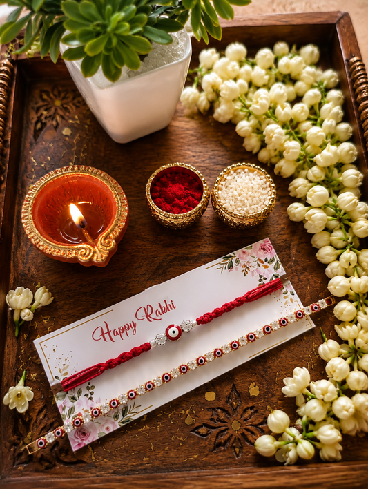 Premium handcrafted Evil Eye Rakhi and matching bracelet combo displayed on a decorative wooden tray with diya, kumkum, rice, and fresh jasmine flowers for Raksha Bandhan celebration.