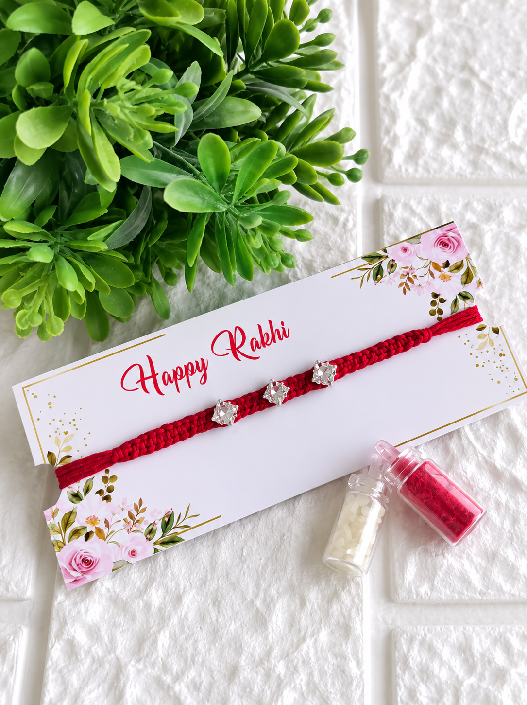 A contemporary Raksha Bandhan Rakhi featuring a thick red braided cord adorned with three square-cut silver solitaire crystal charms, presented on a white floral "Happy Rakhi" card next to two miniature glass vials filled with traditional roli and chawal, placed on a white brick-textured surface under a green leafy plant.