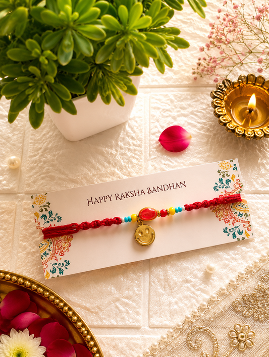 Red braided Rakhi featuring a dangling gold smiley face charm, central oval red stone, and vibrant accent beads on a Happy Raksha Bandhan card.