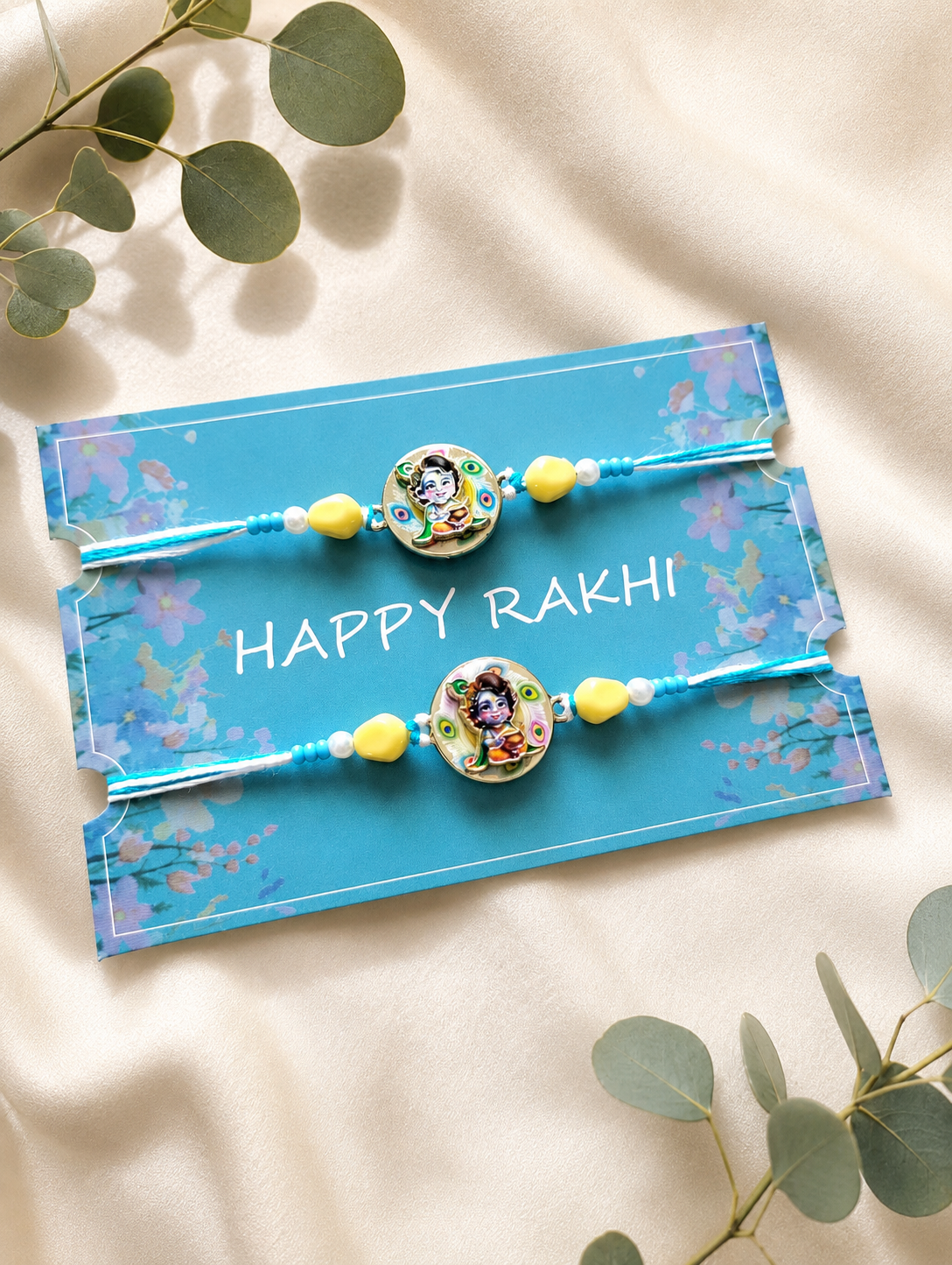 Set of two handcrafted Bal Krishna rakhis with blue and yellow beads, displayed on a blue "Happy Rakhi" card surrounded by green leaves on a beige fabric background.