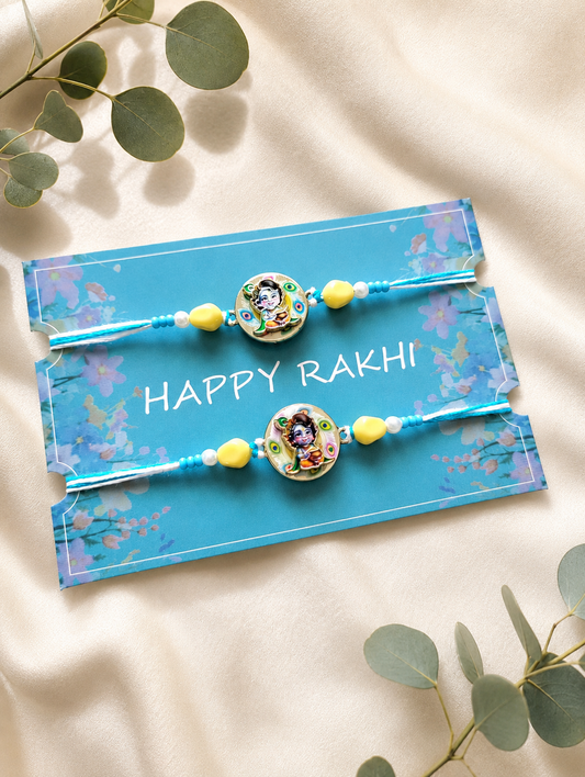 Set of two handcrafted Bal Krishna rakhis with blue and yellow beads, displayed on a blue "Happy Rakhi" card surrounded by green leaves on a beige fabric background.