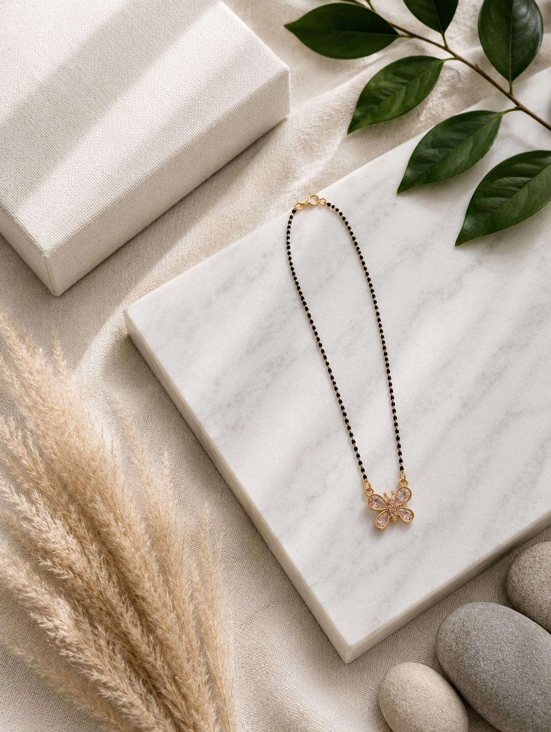 A product photography showcase of a modern gold-toned mangalsutra featuring a butterfly pendant set with clear crystals and a traditional fine chain interspersed with dainty black beads. The collection includes a styled flat-lay layout on a white marble slab surrounded by textured textiles and botanical accents, and a macro close-up view detailing the stone-encrusted butterfly charm.
