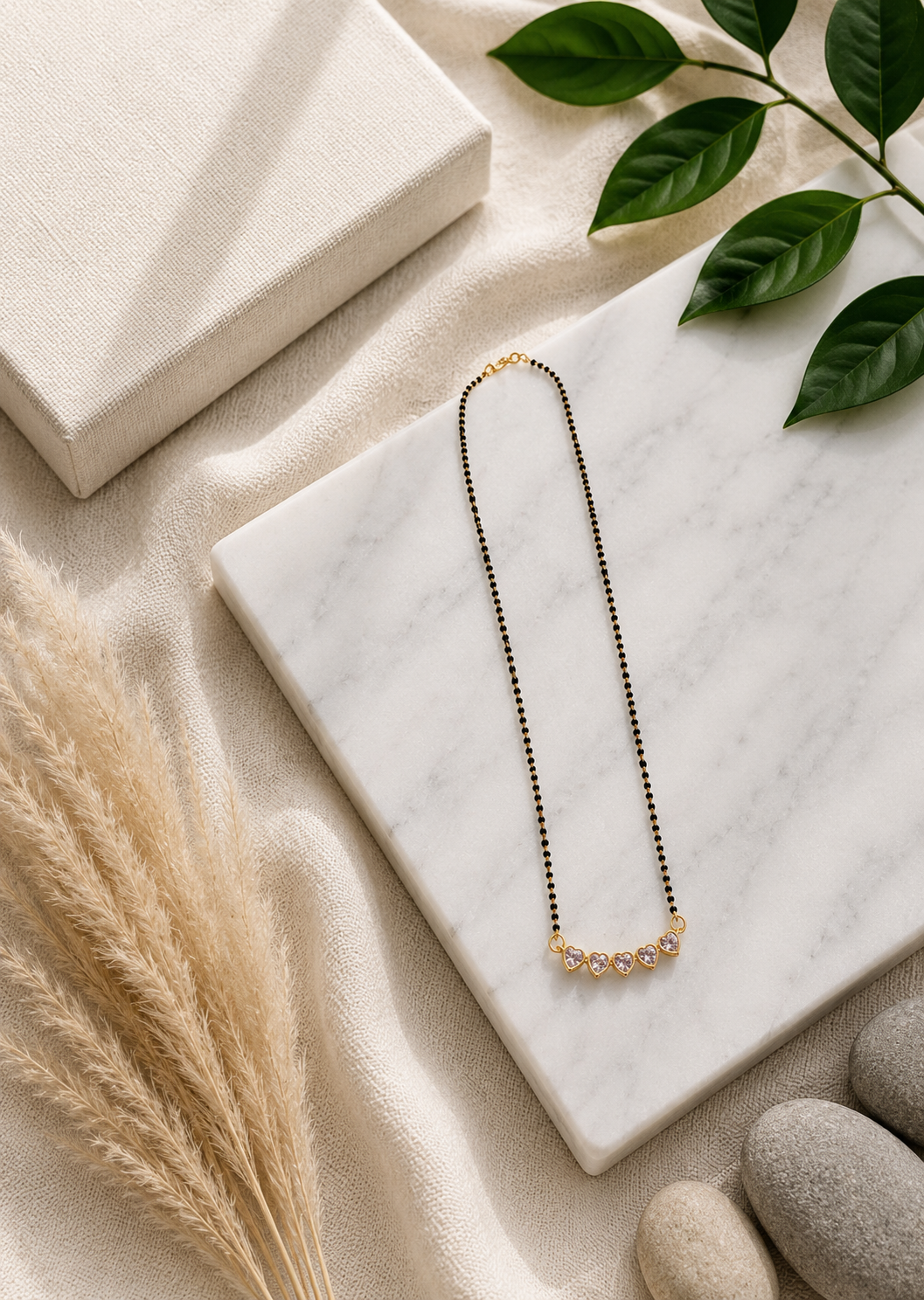 A product photography showcase of a contemporary gold-toned mangalsutra featuring a curved pendant made of five interconnected heart-shaped clear crystals and a delicate chain threaded with small black beads. The collection includes a styled flat-lay view on a white marble block surrounded by soft organic elements, and a close-up detail shot highlighting the brilliant facets of the heart stones and their polished gold bezels.