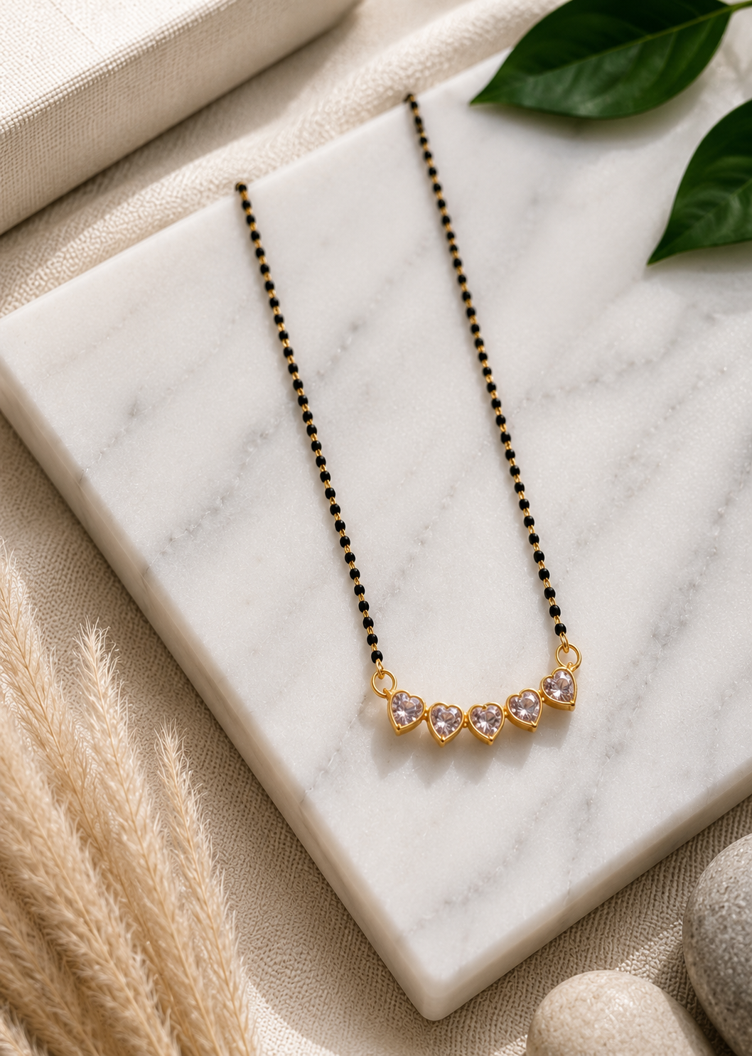A product photography showcase of a contemporary gold-toned mangalsutra featuring a curved pendant made of five interconnected heart-shaped clear crystals and a delicate chain threaded with small black beads. The collection includes a styled flat-lay view on a white marble block surrounded by soft organic elements, and a close-up detail shot highlighting the brilliant facets of the heart stones and their polished gold bezels.