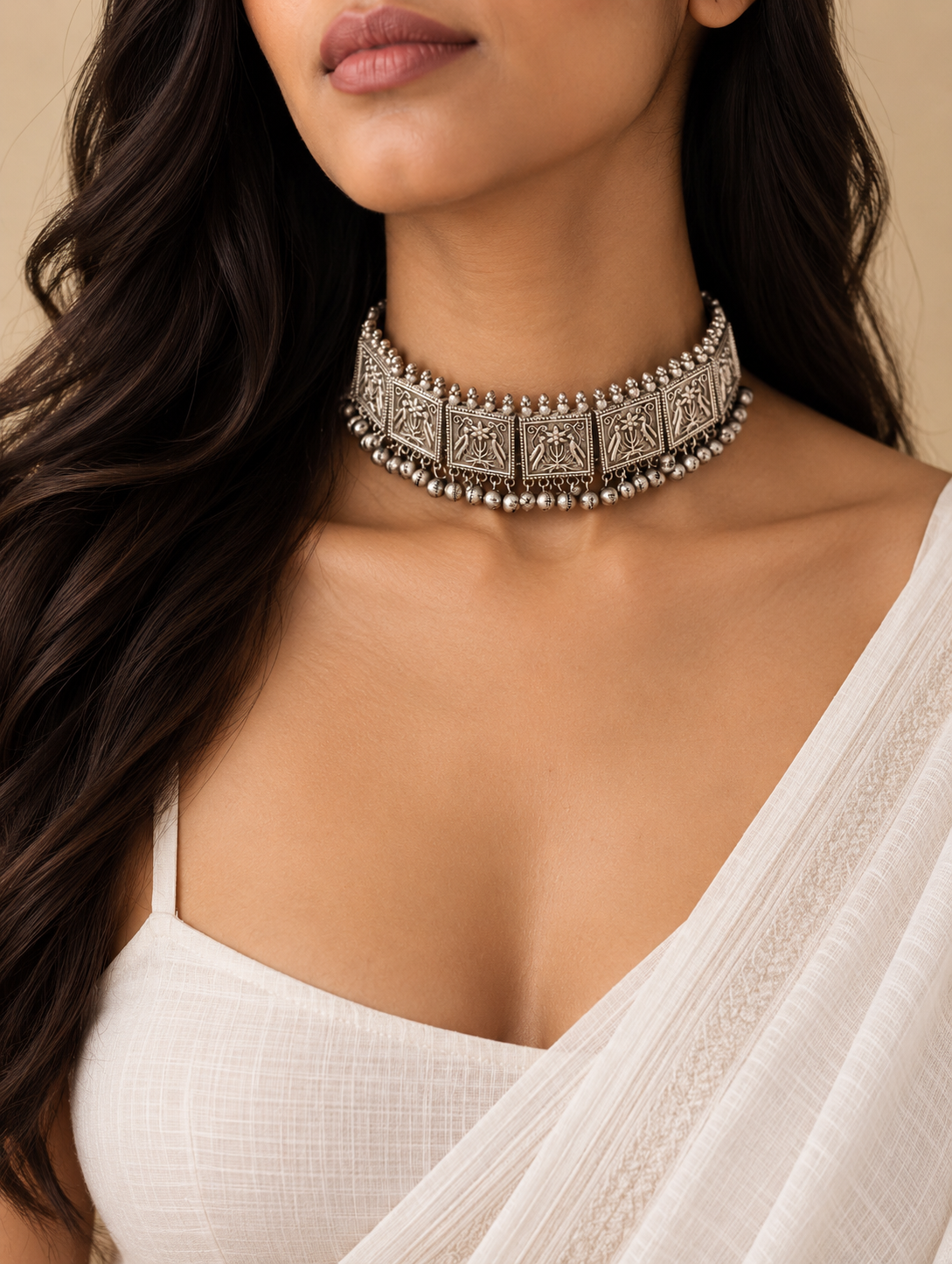 A close-up of a woman wearing a silver-toned, heavily embellished choker necklace with square motifs and dangling bead details, paired with a white draped top against a warm beige background.