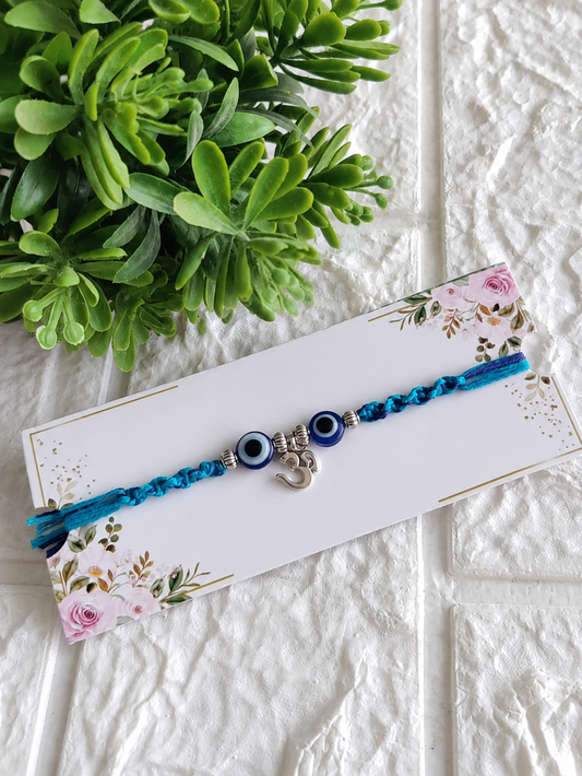 A Raksha Bandhan Rakhi featuring a dual-toned blue and turquoise braided cord, adorned with a central dangling silver Om charm flanked by two blue-and-white Evil Eye beads, displayed on a white floral backing card next to a green potted plant.