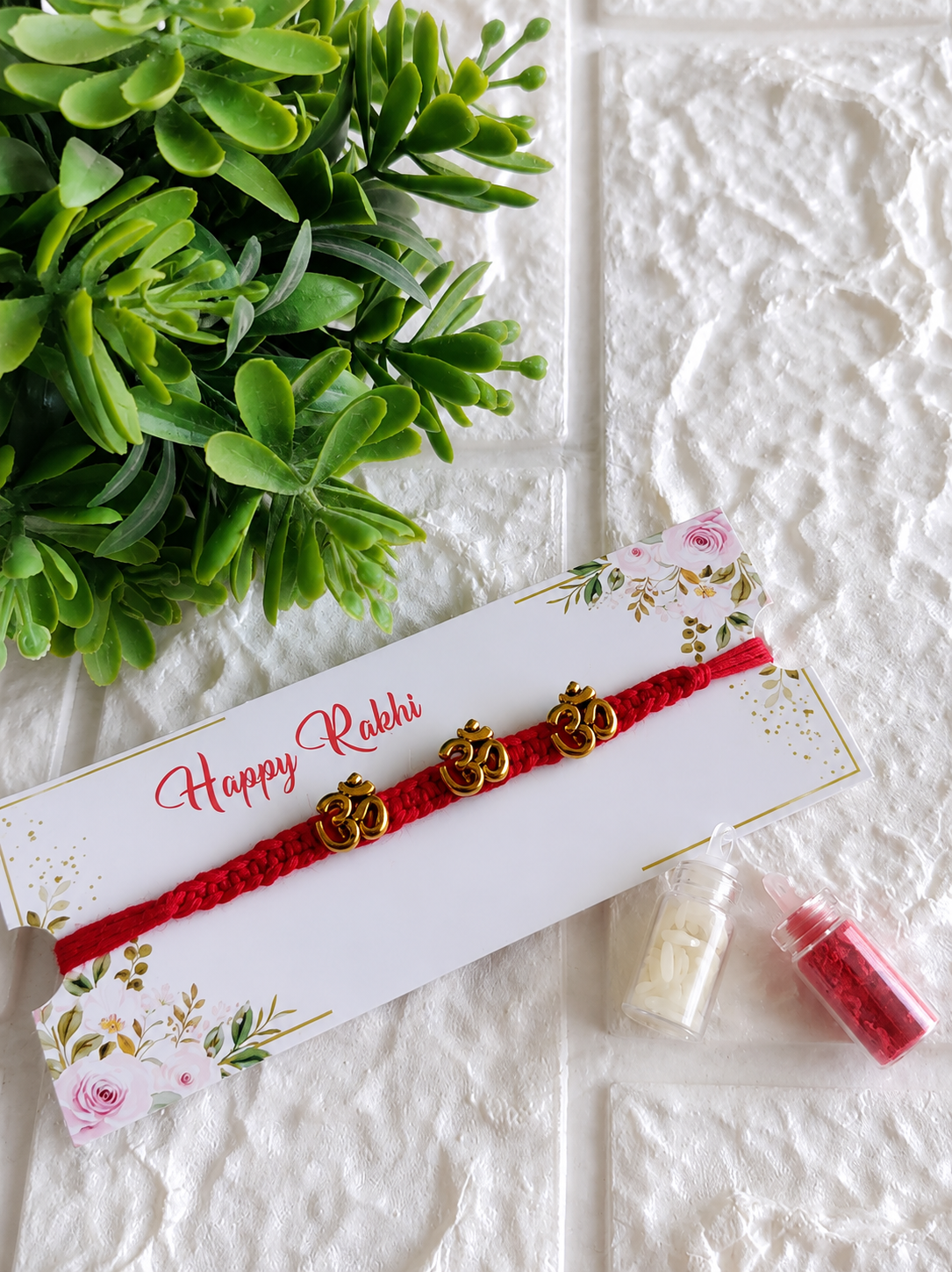 A spiritual Raksha Bandhan Rakhi featuring a thick red braided cord adorned with three horizontally aligned golden "Om" symbols, displayed on a white floral "Happy Rakhi" card next to two miniature glass vials filled with roli and chawal, set against a white textured background with a green plant.