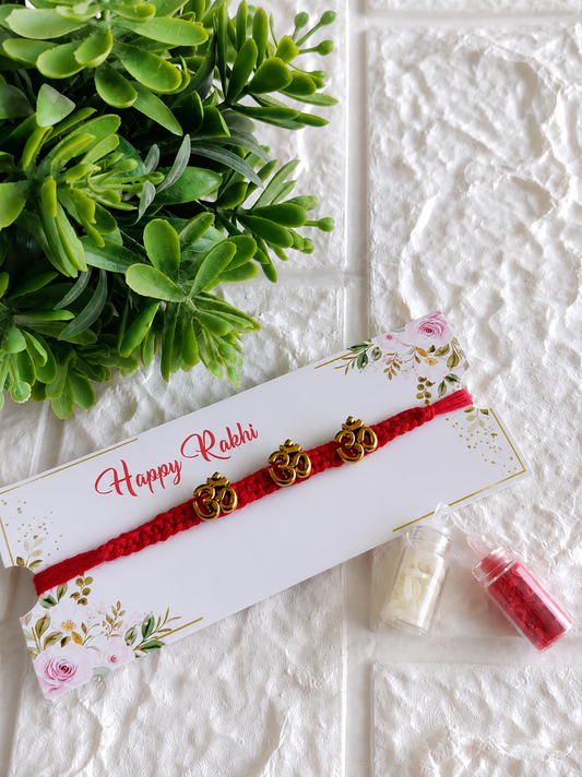 A spiritual Raksha Bandhan Rakhi featuring a thick red braided cord adorned with three horizontally aligned golden "Om" symbols, displayed on a white floral "Happy Rakhi" card next to two miniature glass vials filled with roli and chawal, set against a white textured background with a green plant.