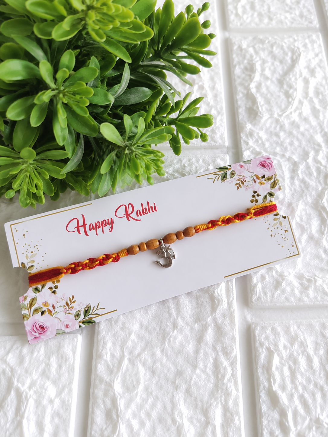 A traditional Raksha Bandhan Rakhi featuring a red and yellow braided cotton cord adorned with a central dangling silver Om charm and six oval wooden beads, displayed on a white floral "Happy Rakhi" card alongside a green potted plant against a white textured brick background.