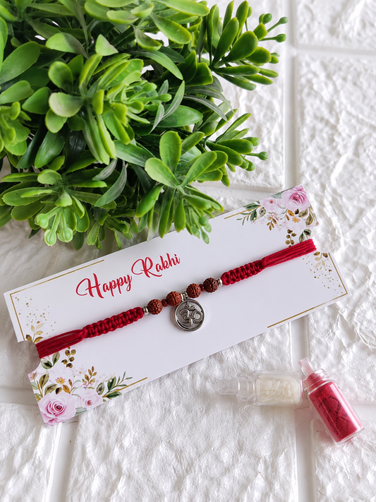 A spiritual Raksha Bandhan Rakhi featuring a thick red braided cotton cord adorned with a central circular silver Om charm and four textured Rudraksha beads, displayed on a white floral "Happy Rakhi" card next to two miniature glass vials filled with roli and chawal, set against a white brick-textured background with a green plant.
