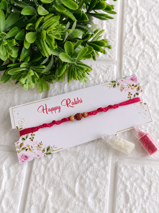 A traditional Raksha Bandhan Rakhi featuring a thick red braided cotton cord adorned with a single central textured Rudraksha bead flanked by two smooth oval wooden beads, displayed on a white floral "Happy Rakhi" card next to two miniature glass vials filled with roli and chawal, set against a white brick-textured background with a green plant.