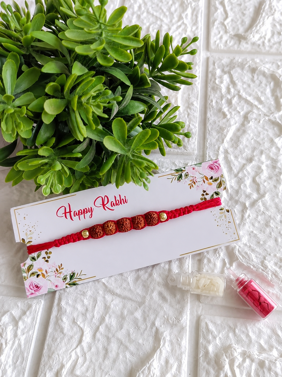 A traditional Raksha Bandhan Rakhi featuring a thick red braided cotton cord adorned with a central row of five textured Rudraksha beads flanked by two small gold beads, displayed on a white floral "Happy Rakhi" card next to two miniature glass vials filled with roli and chawal, set against a white brick-textured background with a green plant.