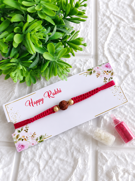 A contemporary Raksha Bandhan Rakhi featuring a thick red braided cotton cord adorned with a single central textured Rudraksha bead flanked by two small gold beads, displayed on a white floral "Happy Rakhi" card next to two miniature glass vials filled with roli and chawal, set against a white brick-textured background with a green plant.