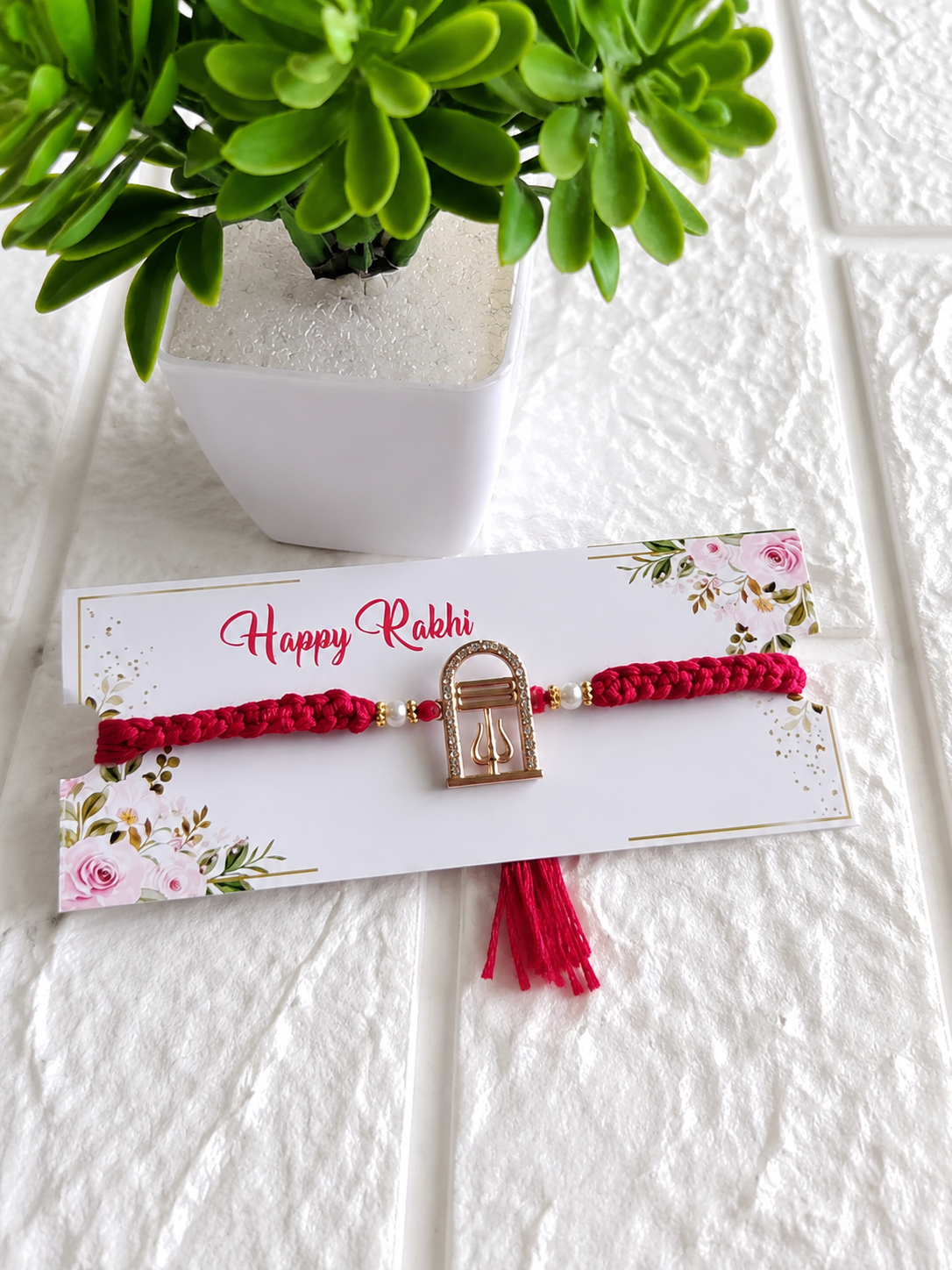 A spiritual Raksha Bandhan Rakhi featuring a thick red braided cotton cord and a central golden charm shaped like an archway containing a Shivling and a Trishul with rhinestone accents, displayed on a white floral "Happy Rakhi" card next to a green potted plant.