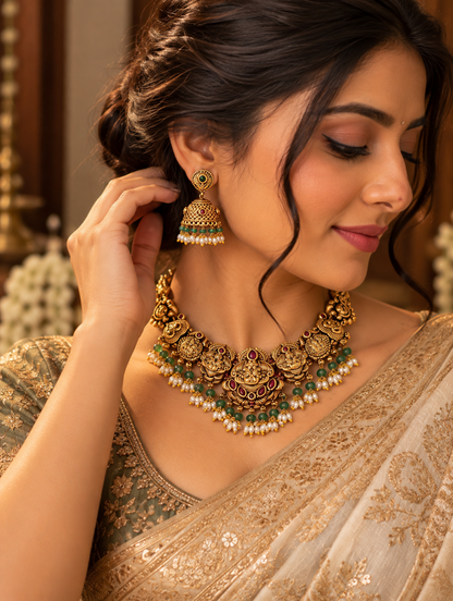 A close-up lifestyle shot of a woman wearing an ornate gold-toned traditional necklace and matching jhumka earrings, styled with a beige and green embroidered saree.